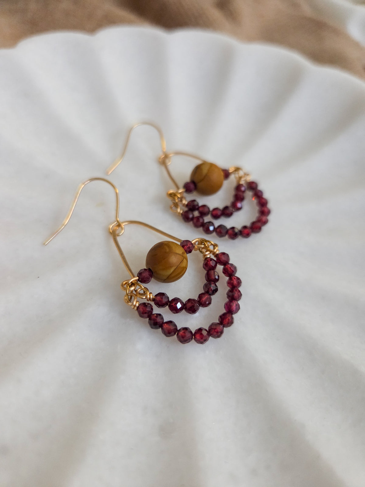 A pair of bohemian-style gold hoop earrings with a large round brown wood jasper bead and a smaller double-strand beaded drop in deep burgundy red garnet, resting on a white scalloped dish.