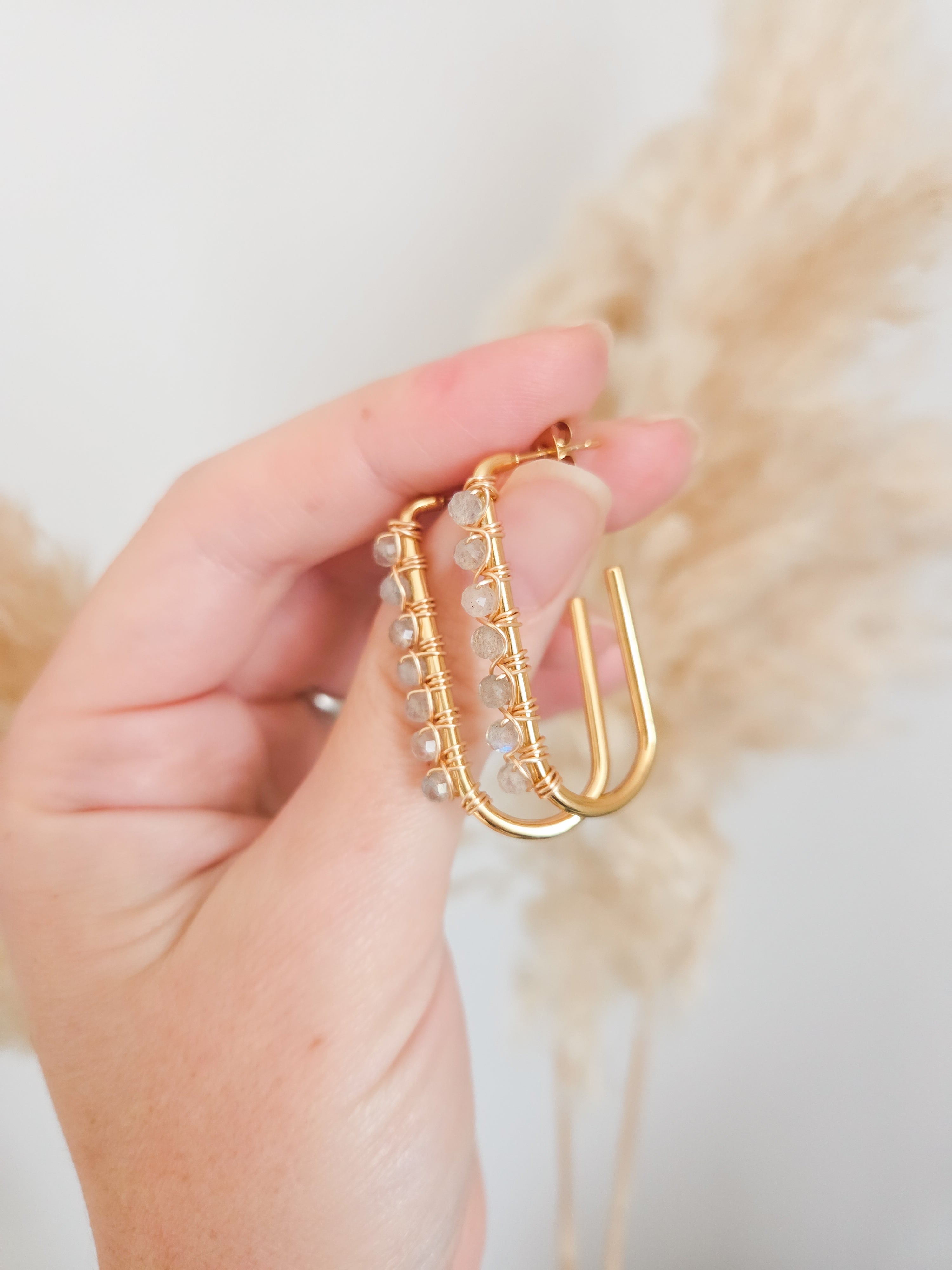 Gold hoop earrings with faceted labradorite gemstone beads held by a hand against a neutral background