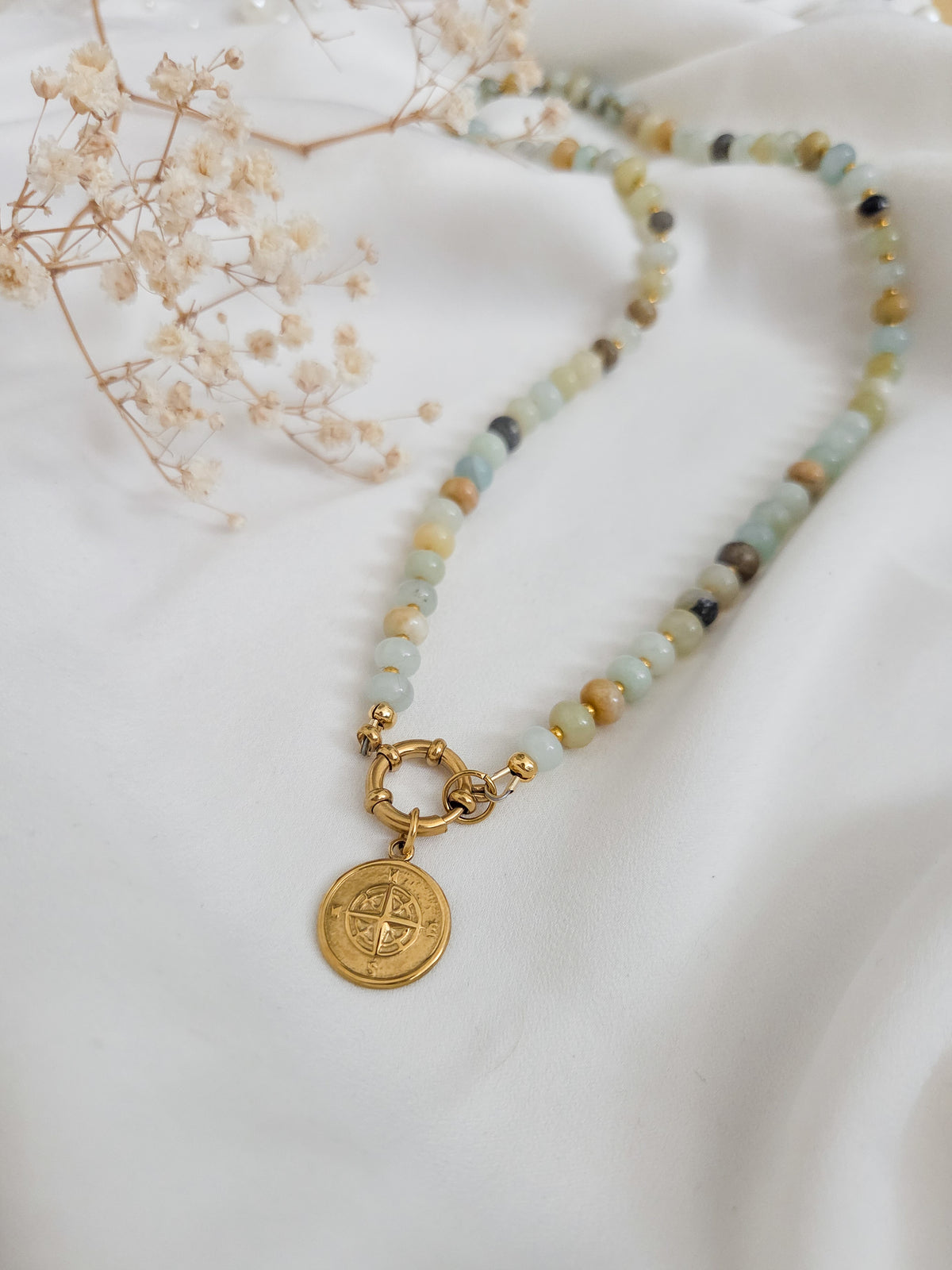 A beaded necklace with light blue, green, and beige stones, featuring a gold clasp and a round gold compass pendant, displayed on a white fabric background with dried baby's breath flowers.