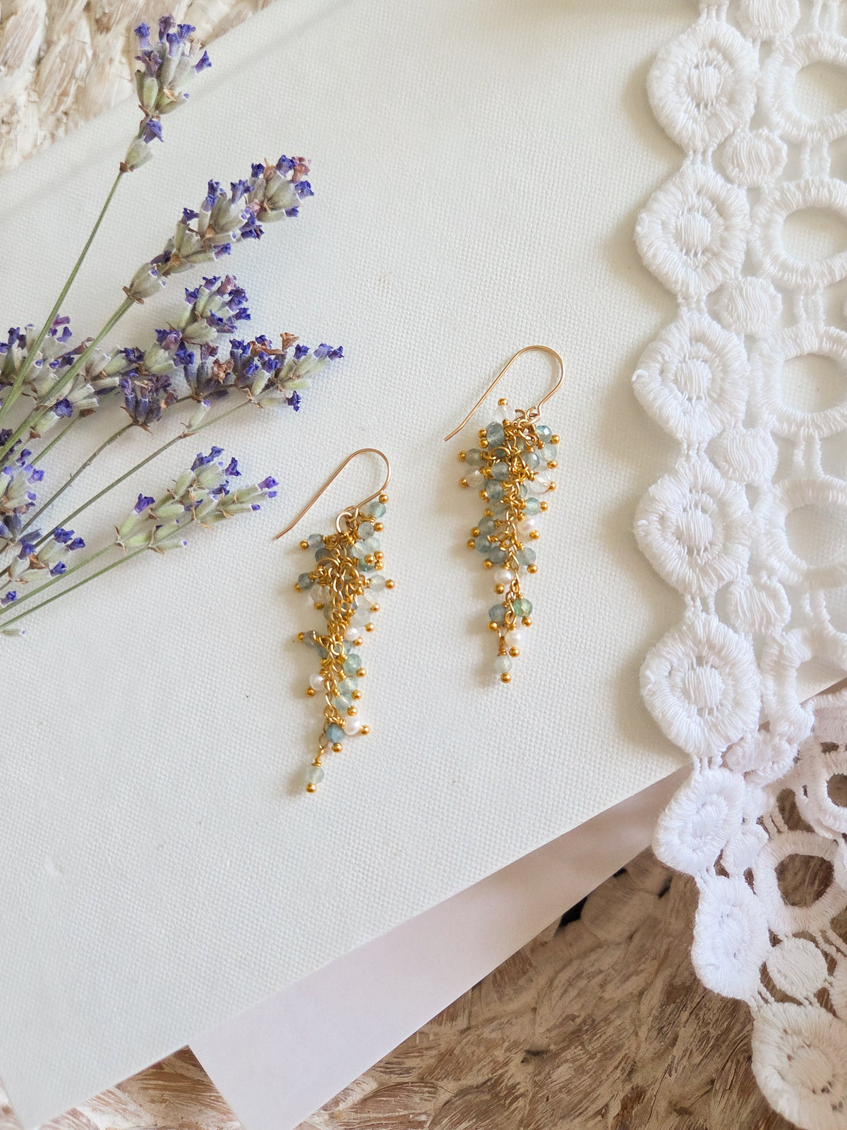 A pair of gold-toned dangle earrings with small pearls and faceted blue-green and clear gemstones, styled on a white card with lavender sprigs, a white lace doily, and a beige knit blanket.