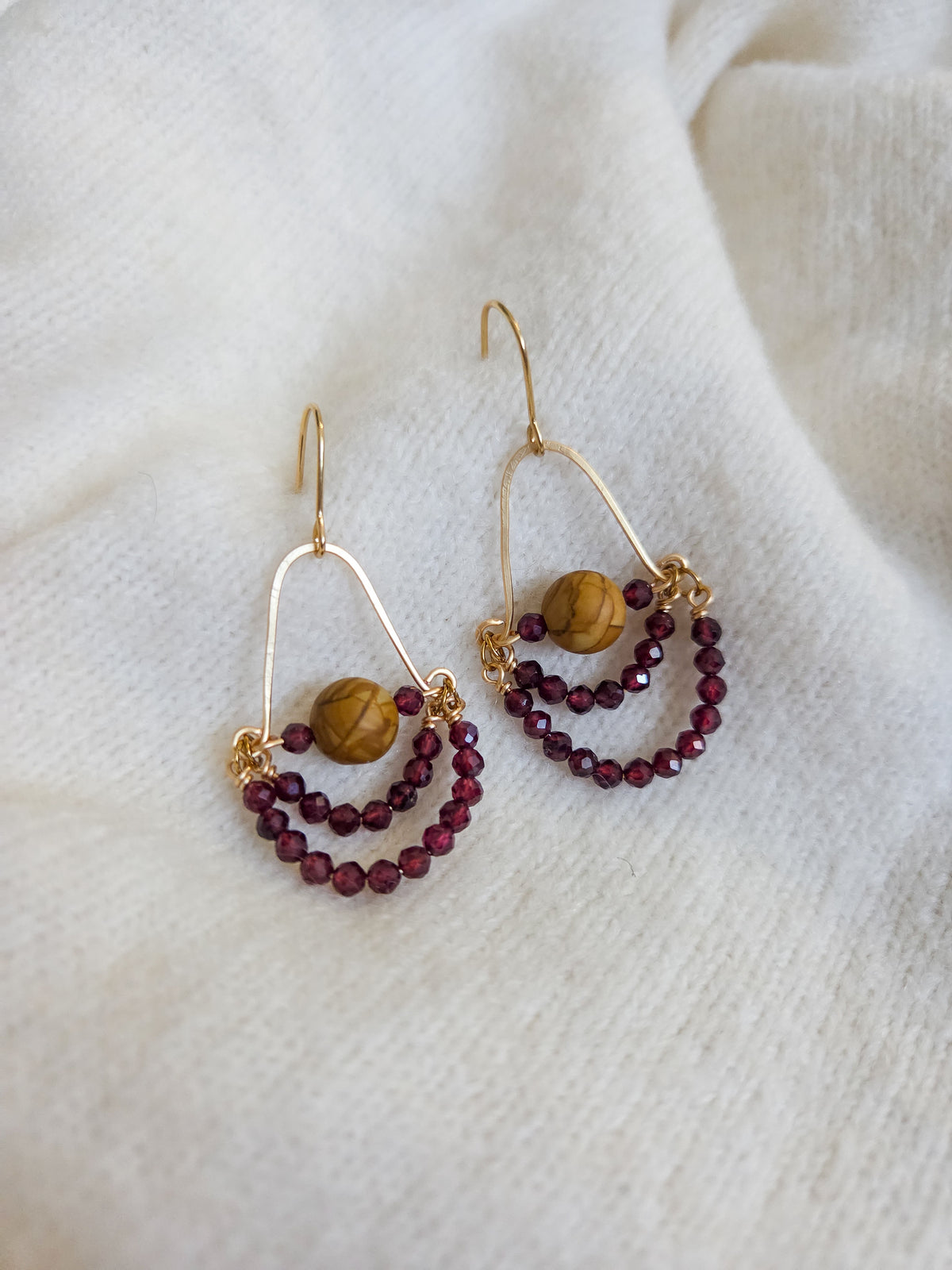 A pair of gold-tone dangle earrings with a large arch, a round brown wooden bead, and two smaller beaded crescent loops in deep burgundy, displayed on a soft white textured fabric background.