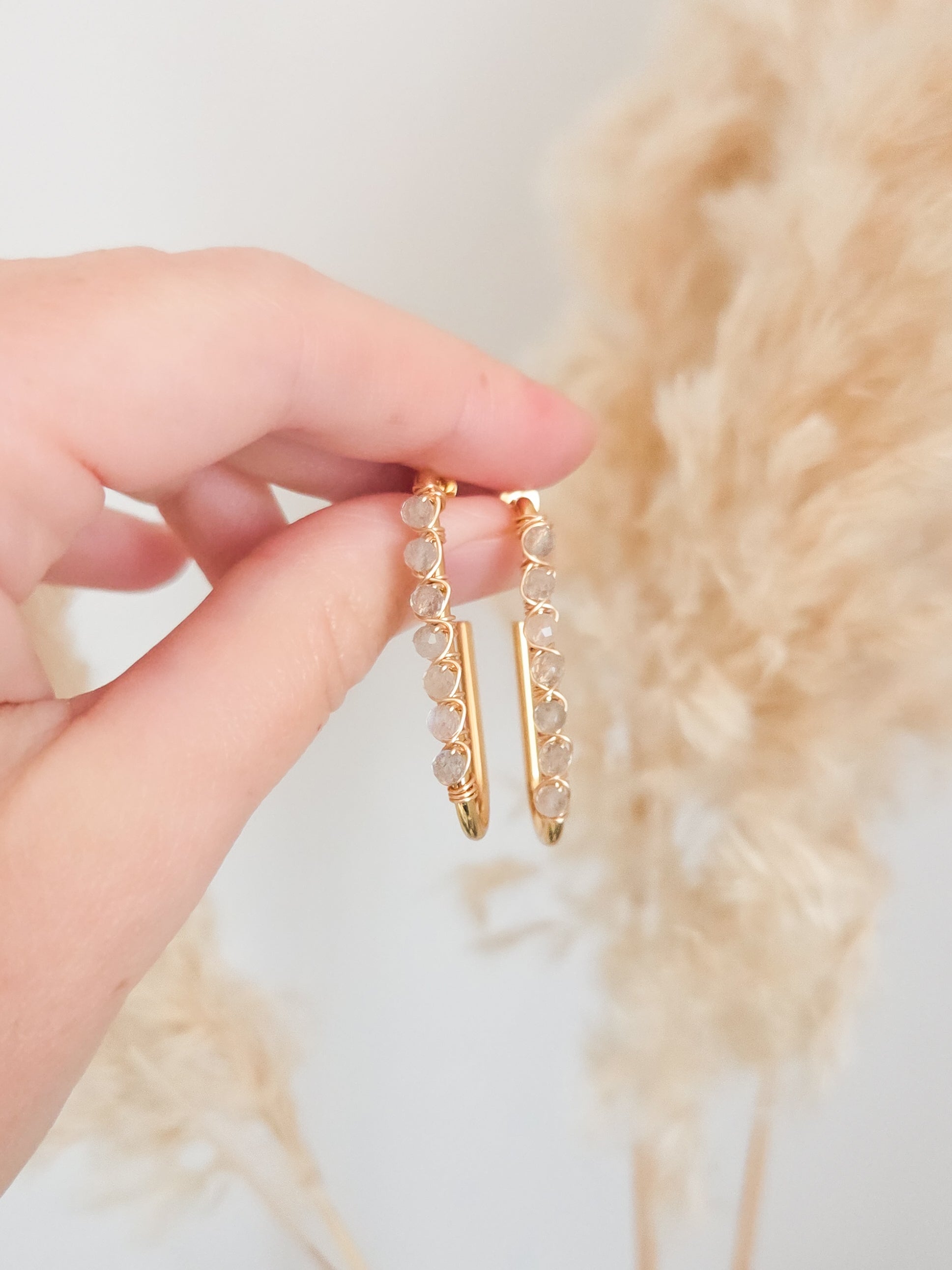 Gold hoop earrings with small stones held by a hand against a soft, blurred background. Neutral gemstone earrings.