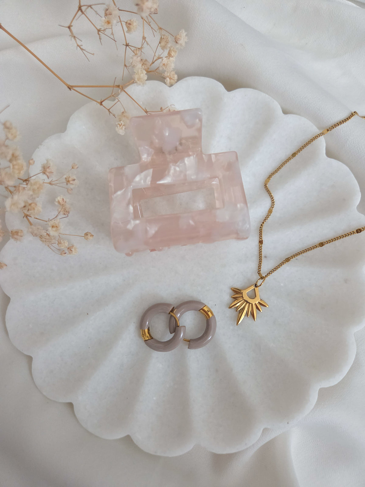 A styled flat-lay of jewelry and a hair accessory on a white scalloped tray with dried flowers. The set includes a translucent blush pink hair claw clip, a gold chain necklace with a sun pendant, and two small gold-and-white hoop earrings.