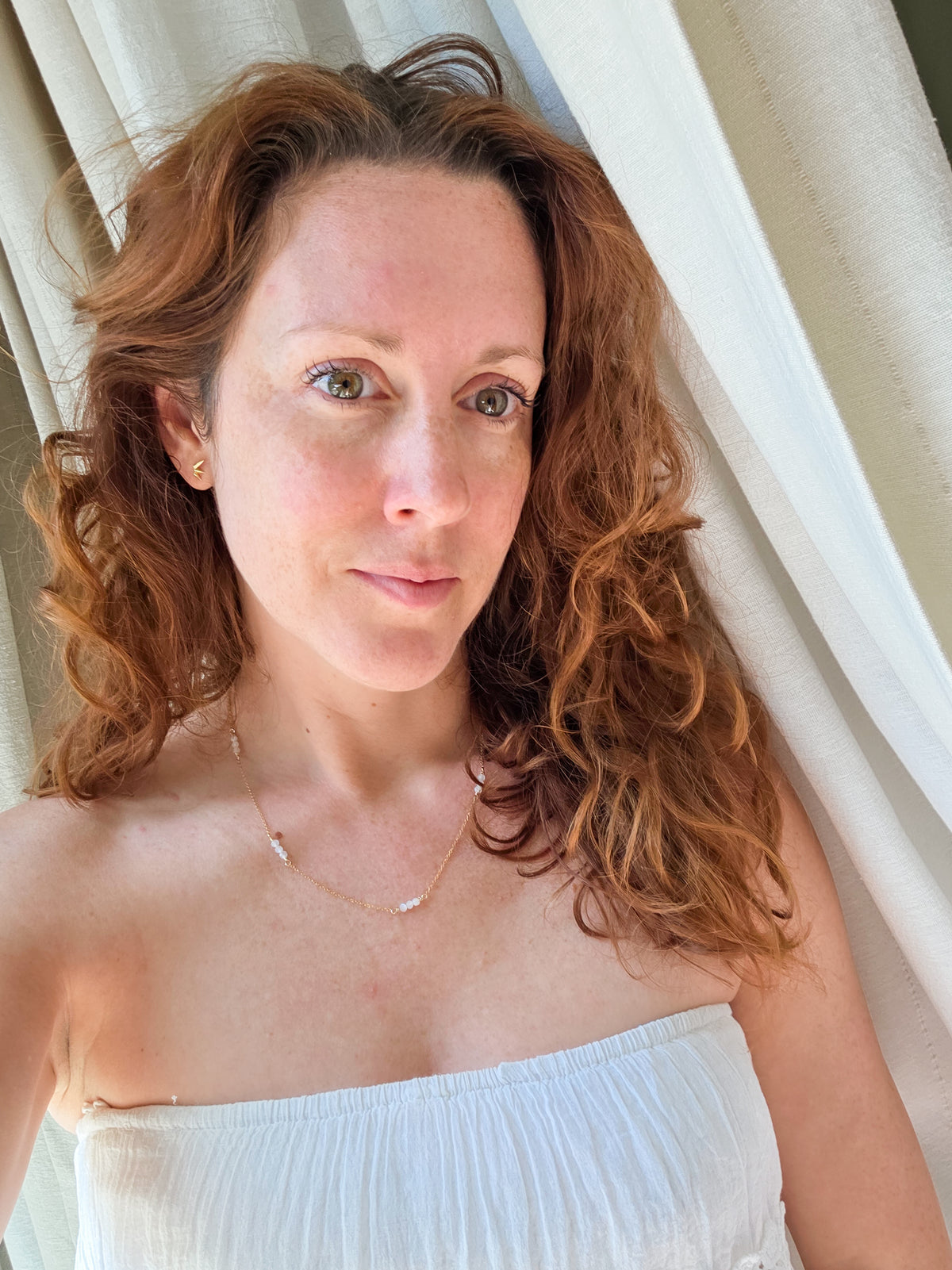 A woman with curly red hair wearing a white strapless top and a delicate gold necklace, posing in front of white curtains.