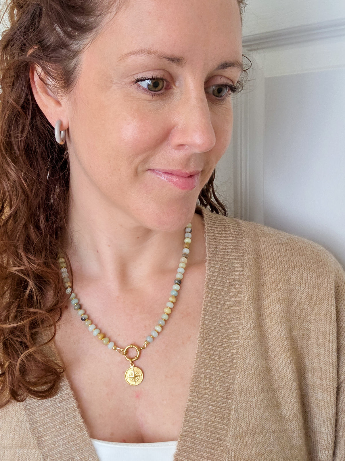 A woman wearing a gold pendant necklace with light green and white beads, styled with a beige cardigan. Best jewelery gifts for women in Canada.