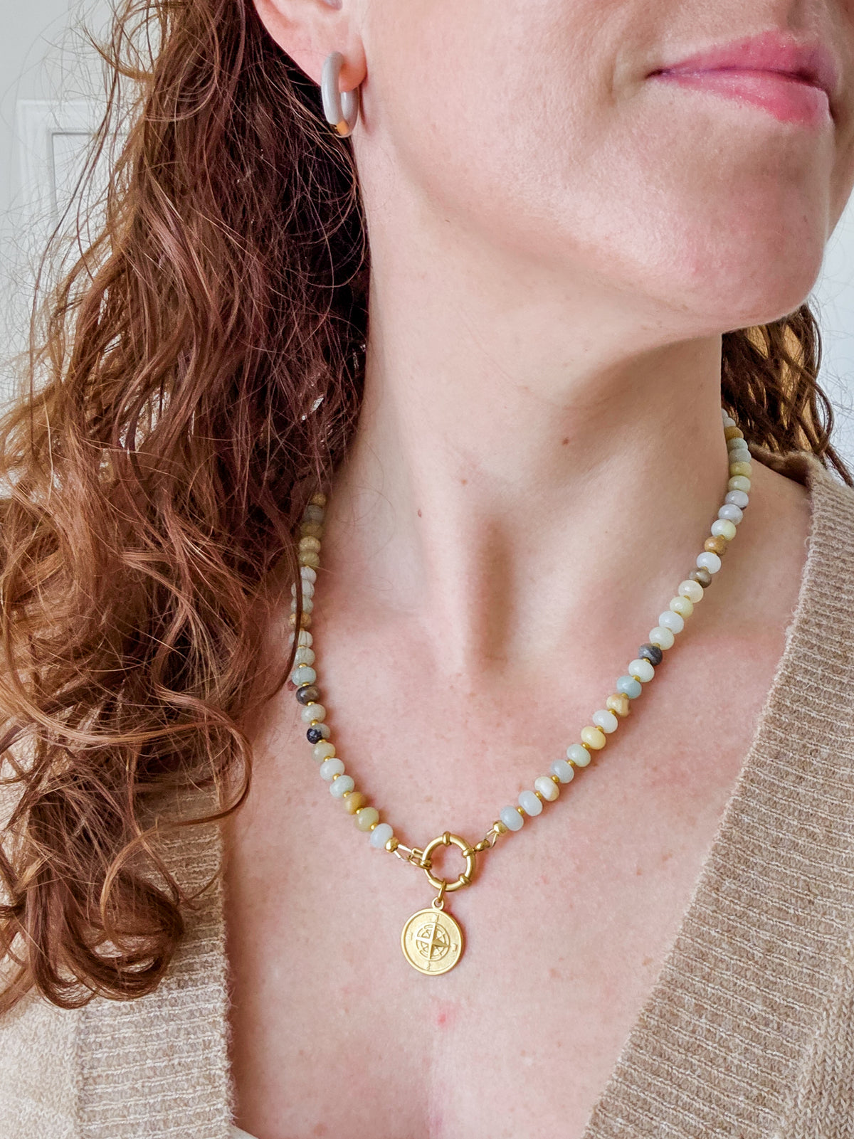 A close-up of a woman wearing a delicate beaded necklace with a gold clasp and a round gold pendant featuring a cross design. The necklace features small, multi-colored beads in shades of cream, light blue, and pale green.