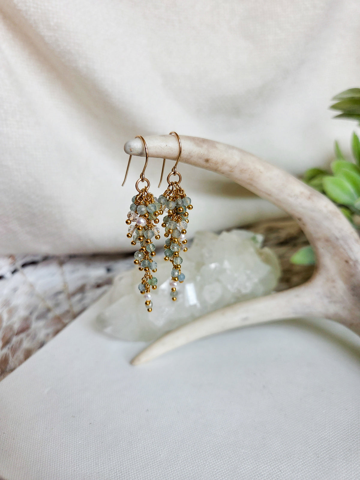 A pair of dangling earrings with gold-toned hooks and clusters of small pale green and white beads, hanging from a decorative antler prop on a soft cream fabric background.