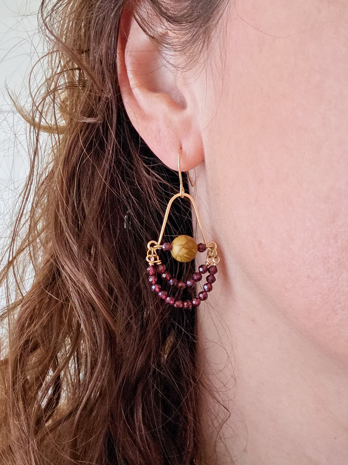 A close-up of a person's ear wearing a gold-tone dangle earring with a large round tan bead and two smaller beaded loops in dark burgundy.