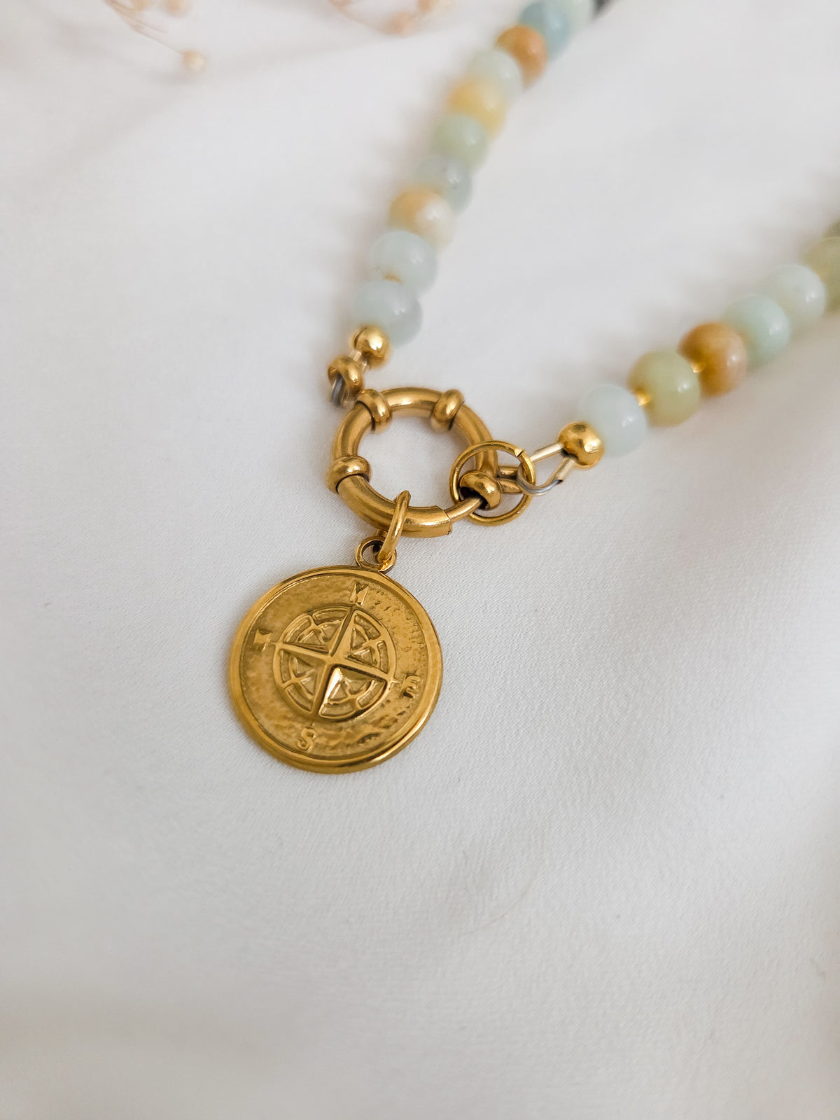 A close-up of a beaded necklace with light green, blue, and tan round beads, featuring a gold-toned toggle clasp and a circular gold pendant with an engraved compass design, resting on a white textured fabric.