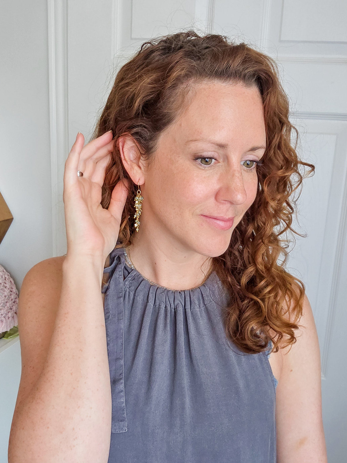 A woman with curly light brown hair wearing a sleeveless slate blue dress and gold dangle earrings, standing in front of a white paneled door.