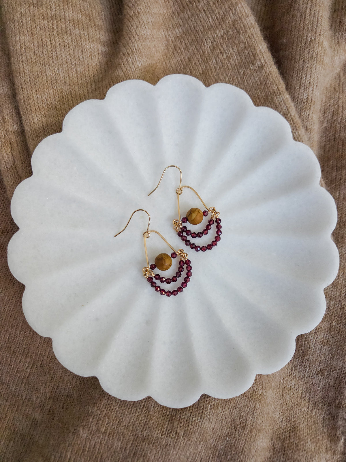 A pair of gold-tone dangle earrings with small dark red garnet beads and a central wood jasper bead, displayed on a white scalloped dish over a brown textured fabric background.