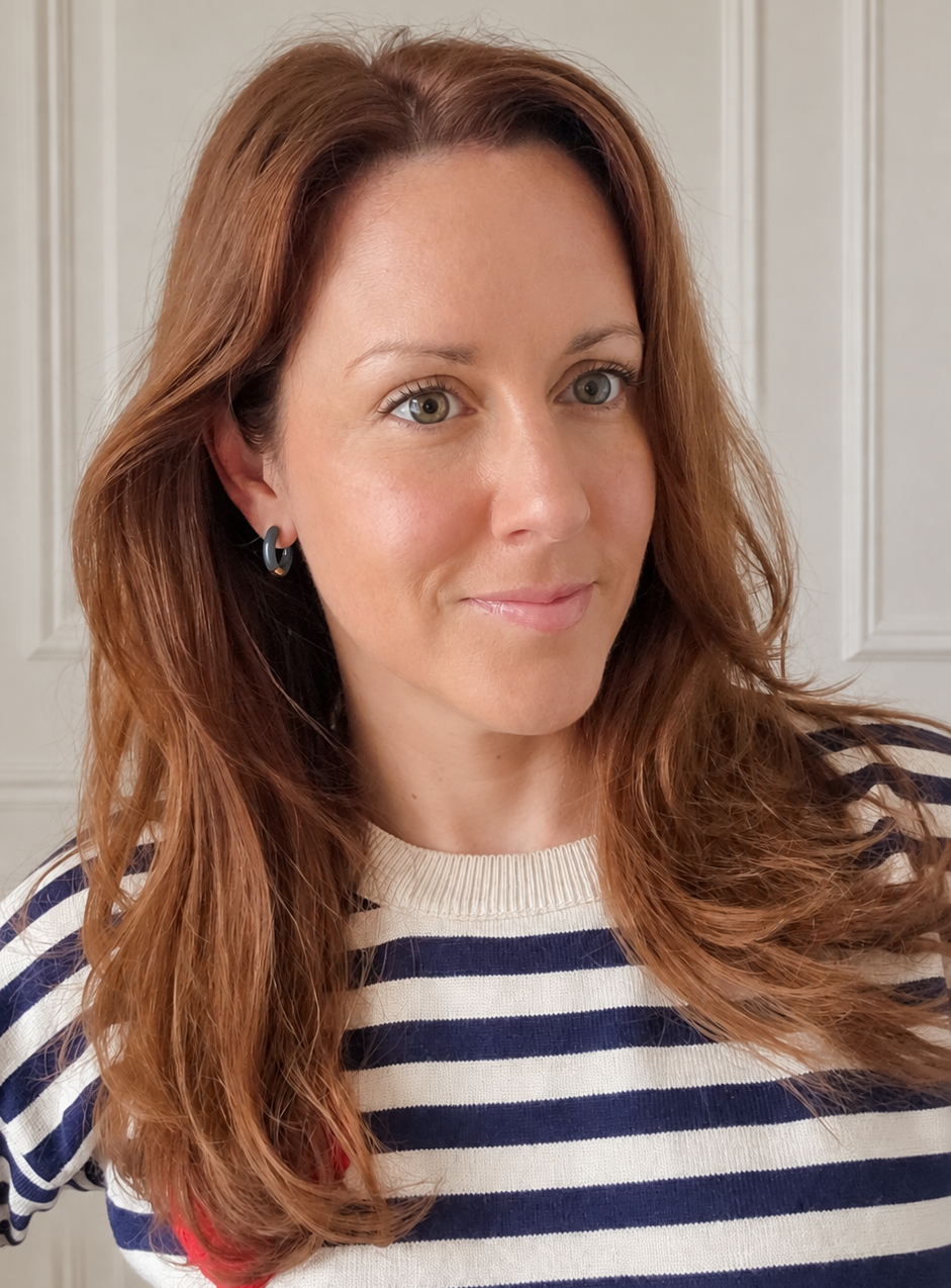 A woman with long auburn hair wearing a navy and white striped knit sweater wearing small teal hoop earrrings, photographed against a light paneled wall.