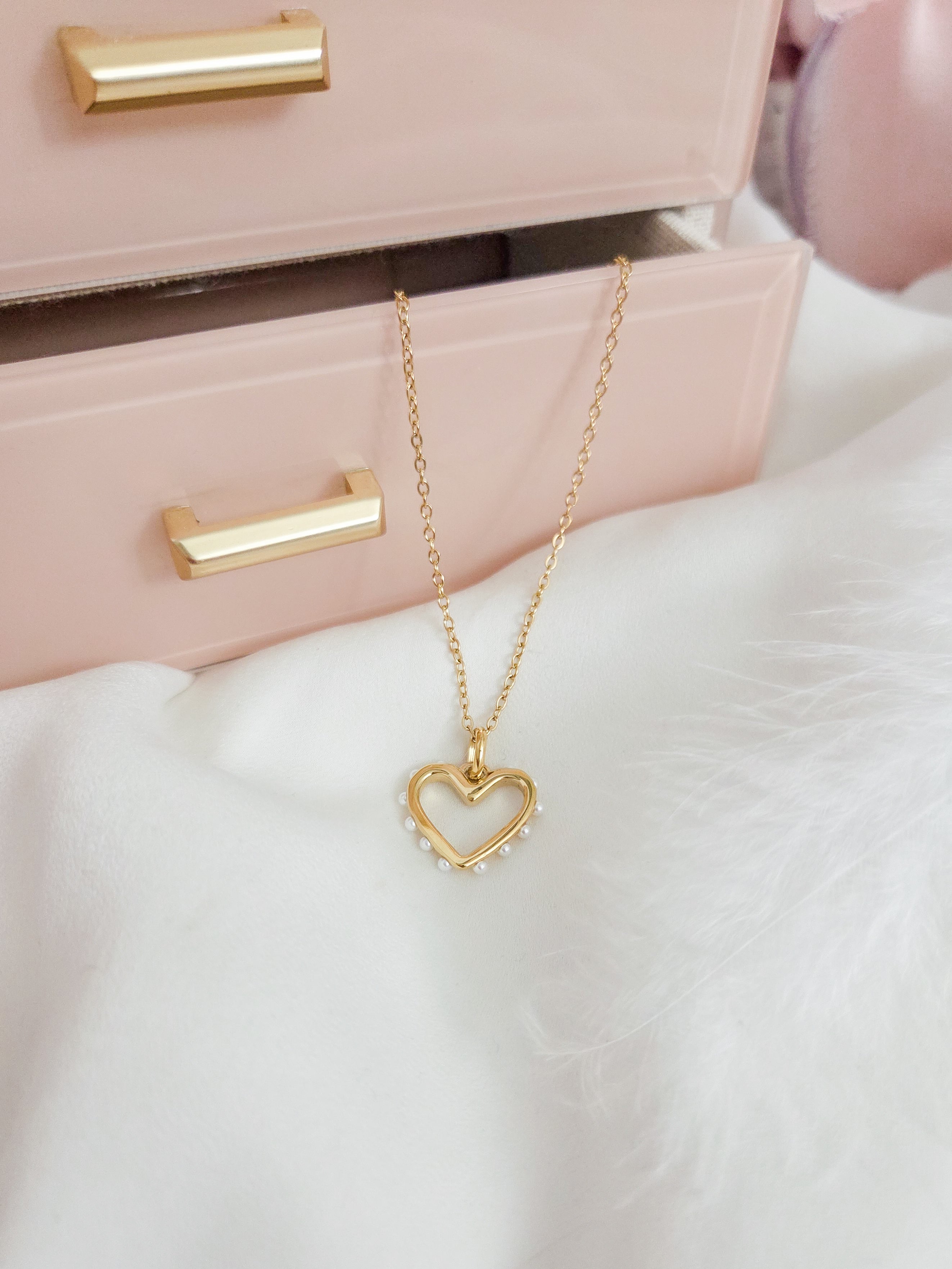 Gold heart-shaped necklace on a white surface with a pink jewelry box in the background.